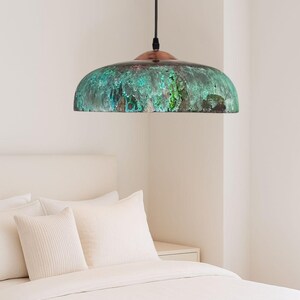 May include: A dome-shaped pendant light with a copper top and a verdigris-coloured shade. The shade has a mottled green and brown appearance. The light hangs from a black cord. The bedroom setting includes a white bed and pillows.