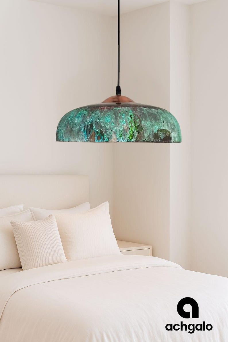 May include: A dome-shaped pendant light with a copper top and a verdigris-coloured shade. The shade has a mottled green and brown appearance. The light hangs from a black cord. The bedroom setting includes a white bed and pillows.