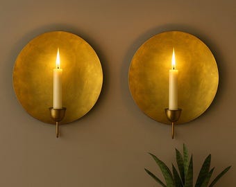 May include: Two wall-mounted candle holders with a gold finish. Each holder features a round, textured gold plate with a small candle cup and a lit, white candle. The background is a neutral brown color, and a green plant is visible.