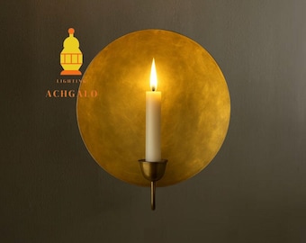 May include: A wall-mounted candle holder with a lit white candle. The candle is set against a large, round, gold-colored metal disc. The holder is made of brass. The logo "ACHGALO LIGHTING" is in the upper left corner.