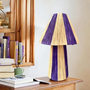 May include: A purple and natural woven lamp with a mushroom-shaped base and a wide, striped lampshade. The lamp is sitting on a wooden table.