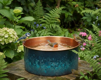 May include: A round bird bath with a copper-coloured interior and a textured, teal-patina exterior. Three small birds are perched on the rim and inside the water-filled basin, which is set upon a stone surface surrounded by lush green foliage.