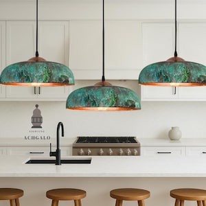 May include: Three pendant lights with verdigris shades and copper rims hang above a kitchen island. The kitchen features white cabinets, a black tap, and wooden stools. The logo "ACHGALO" is visible on the wall.