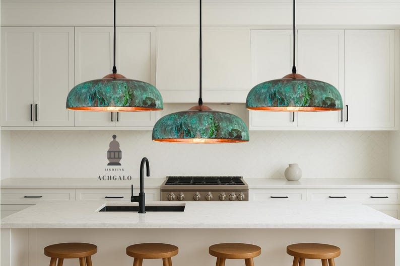 May include: Three pendant lights with verdigris shades and copper rims hang above a kitchen island. The kitchen features white cabinets, a black tap, and wooden stools. The logo "ACHGALO" is visible on the wall.