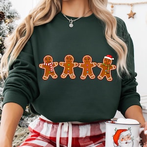 Op de afbeelding: Donkergroene sweatshirt met vier peperkoekenmannetjes. Elk peperkoekenmannetje heeft een unieke sjaal en knoopontwerp. Eén peperkoekenmannetje draagt een kerstmuts. Een feestelijk vakantieontwerp.