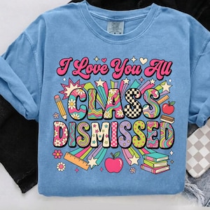 Puede incluir: Camiseta azul claro con el texto "I Love You All CLASS DISMISSED" en un diseño retro colorido. El gráfico incluye estrellas, flores, lápices, manzanas y libros. La camiseta está hecha de un material suave.