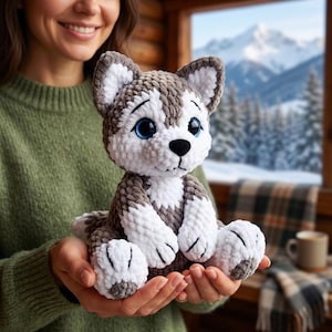 Op de afbeelding: Een gehaakt husky-knuffeldier van zacht grijs en wit garen met grote, expressieve blauwe ogen, een zwarte neus en puntige oren. Het knuffeldier heeft een ronde kop, een witte borst en witte pootjes met grijze accenten in een textuurstijl.
