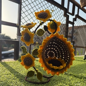 May include: A handcrafted, crochet sunflower sculpture with yellow petals and brown centers. The sculpture features multiple sunflowers of varying sizes, green leaves, and a dark brown metal frame. The sunflowers are arranged to create a decorative piece.