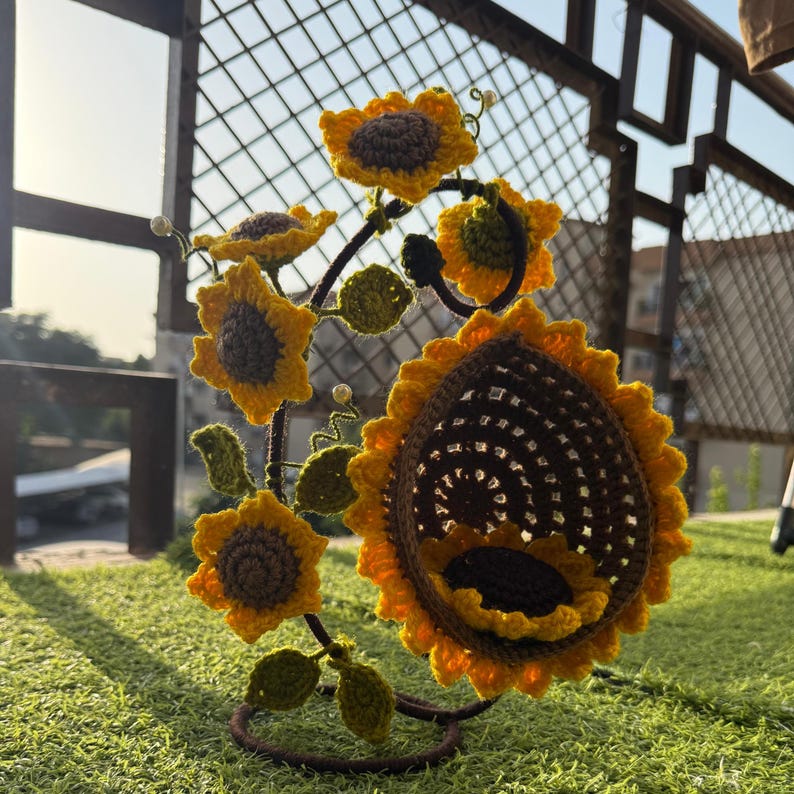 May include: A handcrafted, crochet sunflower sculpture with yellow petals and brown centers. The sculpture features multiple sunflowers of varying sizes, green leaves, and a dark brown metal frame. The sunflowers are arranged to create a decorative piece.