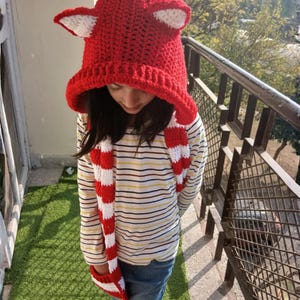 Puede incluir: Un gorro rojo tejido a ganchillo con detalles de orejas blancas y una bufanda a rayas rojas y blancas a juego. El gorro tiene forma de zorro. La persona lleva una camisa a rayas y vaqueros azules. El gorro y la bufanda están hechos a mano.