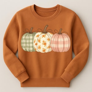 以下が含まれることがあります： 3つの装飾的なカボチャが描かれた、オレンジ色のスウェットシャツ。カボチャは一列に並んでおり、それぞれ異なる模様です。緑のチェック柄、花柄、ピンクのチェック柄。カボチャには茶色の茎があり、ひもで結ばれています。