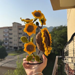 May include: A handcrafted, crochet sunflower hair accessory. The accessory features multiple yellow sunflowers with brown centers and green leaves, attached to a dark brown metal frame. The design is held in a hand against a blurred outdoor background.