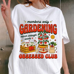 Puede incluir: Camiseta blanca con un diseño temático de jardinería. El gráfico incluye las palabras "members only GARDENING obsessed club" en naranja y verde, junto con ilustraciones de plantas, semillas y herramientas de jardinería.