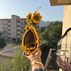 May include: A handmade crochet sunflower-themed birdhouse. The birdhouse is brown and yellow, with a sunflower design. The birdhouse is hanging outside, with a background of buildings and trees.