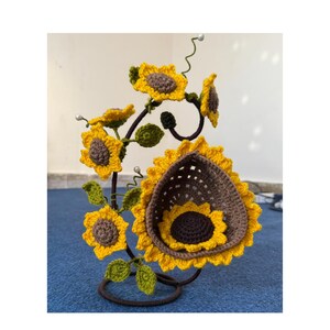 May include: A handcrafted, decorative metal stand featuring crocheted sunflowers. The sunflowers are yellow with brown centers and green leaves. The stand is dark brown and has a circular base. The sunflowers are arranged in a natural, artistic display.