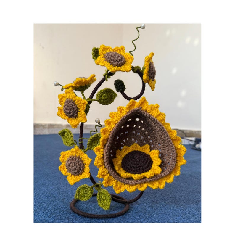 May include: A handcrafted, decorative metal stand featuring crocheted sunflowers. The sunflowers are yellow with brown centers and green leaves. The stand is dark brown and has a circular base. The sunflowers are arranged in a natural, artistic display.