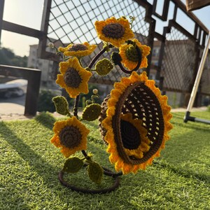 May include: A handcrafted crochet sunflower sculpture with yellow petals and brown centers. The sculpture features multiple sunflowers of varying sizes, green leaves, and a brown metal base. The sunflowers are arranged to create a decorative piece.