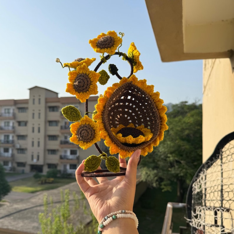 May include: A handcrafted crochet sunflower sculpture with yellow petals and brown centers. The sculpture features multiple sunflowers and green leaves, all attached to a dark metal frame. The sculpture is held up against a blue sky.