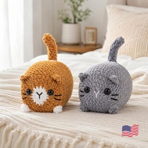 May include: Two plush cat toys made of chunky yarn. One cat is golden-brown with a white patch on its face and white paws, while the other is solid gray. Both feature upright tails, small ears, black plastic eyes, and embroidered whiskers.