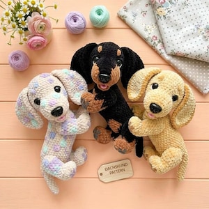 Op de afbeelding: Drie gehaakte teckel knuffels gemaakt van zacht garen. Eén is wit met pastelpaarse en blauwe vlekken, één is zwart met bruine accenten en één is effen bruin. Een klein houten label met de tekst DACHSHUND PATTERN ligt tussen de knuffels in.