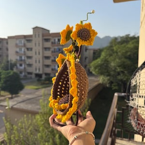 May include: A handmade crochet sunflower-themed hanging basket. The basket is brown with a yellow scalloped edge, and is adorned with yellow and brown crochet sunflowers. The basket is suspended from a green stem with a pearl at the top.