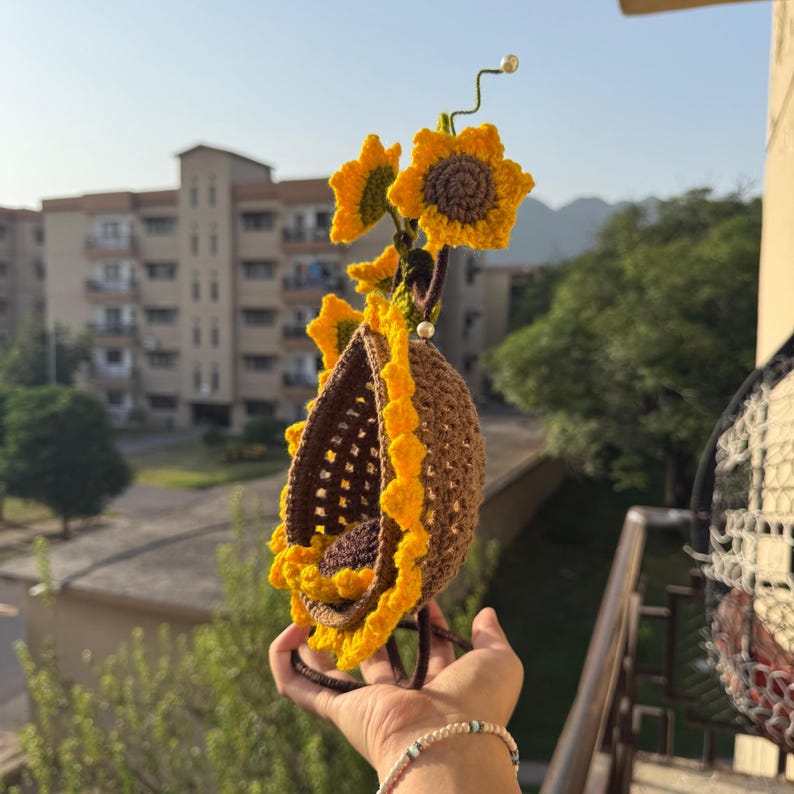 May include: A handmade crochet sunflower-themed hanging basket. The basket is brown with a yellow scalloped edge, and is adorned with yellow and brown crochet sunflowers. The basket is suspended from a green stem with a pearl at the top.