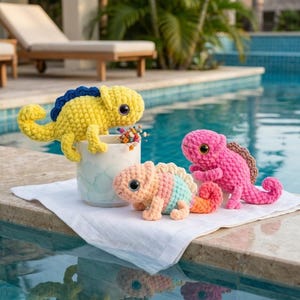 May include: Three crocheted chameleon plush toys. One yellow chameleon with a dark blue crest sits on a white ceramic mug. A smaller peach, teal, and orange striped chameleon sits next to a bright pink chameleon with a brown crest on a white cloth.