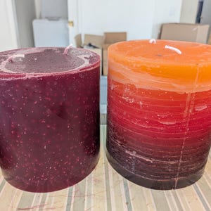 May include: Two cylindrical pillar candles. The left candle is a solid deep burgundy with a textured, speckled surface. The right candle features a layered gradient design transitioning from orange at the top to red and dark purple at the base.