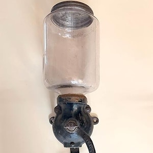 May include: Antique wall-mounted coffee grinder with a clear glass jar, a dark metal body, and a wooden handle. The grinder is attached to a wall with a metal bracket. A vintage kitchen decor item.