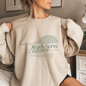Könnte beinhalten: Beiges Sweatshirt mit grünem Tennisschläger-Motiv und den Worten "Work Serve Balance" in Schreibschrift. Der Text "Tennis Club" und "Match Day" sind ebenfalls sichtbar. Das Sweatshirt hat einen Rundhalsausschnitt und lange Ärmel.