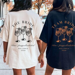 May include: Two oversized t-shirts, one cream and one navy, featuring a graphic of cocktail glasses with orange slices. The cream shirt reads "THE BRIDE" and the navy shirt reads "TEAM BRIDE". Both shirts include text: "Olivia's Junggesellenabschied MALLORCA 2025".