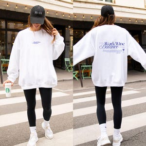 Könnte beinhalten: Ein weißes Sweatshirt mit Rundhalsausschnitt und blauem Text auf der Vorder- und Rückseite. Auf der Vorderseite steht Work Wine Balance. Die Rückseite zeigt denselben Text in größerer Schrift sowie eine kleine Weinglasgrafik und weiteren Text.