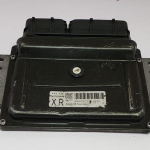 Może przedstawiać: Czarna elektroniczna jednostka sterująca (ECU) o prostokątnym kształcie. ECU ma etykietę z tekstem i kodem kreskowym. Posiada metalowe wsporniki montażowe po bokach i na górze. Na górze znajdują się dwa czarne złącza.