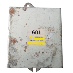 Puede incluir: Un componente electrónico rectangular gris metálico con manchas de óxido. Una etiqueta amarilla muestra el número "601" y otros códigos. El componente tiene una pestaña de montaje y dos pestañas inferiores.