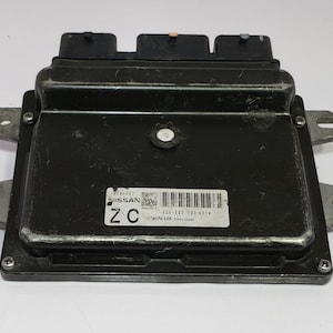 May include: A black electronic control unit (ECU) with a rectangular shape and multiple connectors. The ECU has a label with the Nissan logo and a barcode. The unit is mounted with metal brackets and screws.