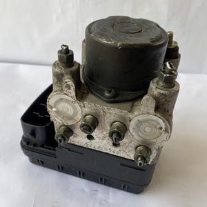 Op de afbeelding: Een gebruikte ABS-module (antiblokkeersysteem). De unit is een combinatie van zwart kunststof en metalen componenten, met meerdere poorten en fittingen. Het bovenste gedeelte is een donkergrijze cilinder. De unit heeft een rechthoekige zwarte basis.