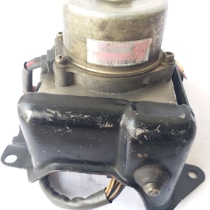 Op de afbeelding: Een gebruikt auto-onderdeel, waarschijnlijk een elektromotor of pomp, met een cilindrische metalen behuizing en een zwarte, rechthoekige basis. Het onderdeel heeft een label met tekst en is gemonteerd op een metalen beugel. Draden en connectoren zijn zichtbaar.