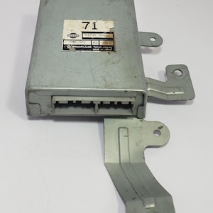 May include: A silver-colored automotive electronic control unit with a rectangular body and an attached bracket. The unit has a label with the number "71" and text including "NISSAN" and "Hitachi, Ltd. Tokyo Japan".