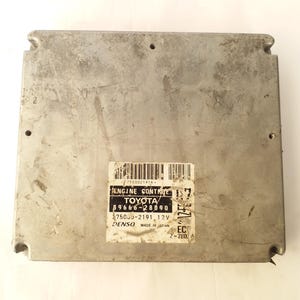 May include: A silver engine control module, rectangular in shape. The label reads "ENGINE CONTROL TOYOTA" along with technical specifications. The module is made in Japan. Dimensions are approximately 20 cm x 20 cm.