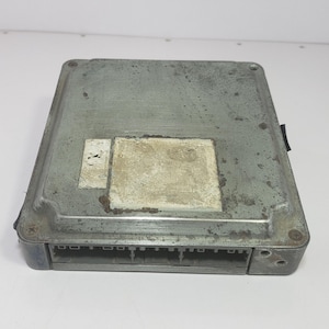 May include: A rectangular, silver-coloured electronic control unit (ECU) with a series of connectors on one side. The unit has a slightly weathered appearance with a rectangular label and a smaller, square label.