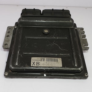 May include: A black Nissan engine control unit (ECU). The unit has a rectangular shape with a series of connectors and a label that reads "NISSAN" and "XB". The ECU is designed for automotive applications.