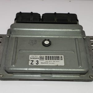 Op de afbeelding: Een zilverkleurige Nissan motorregeleenheid (ECU) met zwarte kunststof componenten. De ECU heeft een label met het Nissan-logo, een barcode en de tekst "Z 3". De ECU is rechthoekig met montagebeugels.