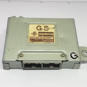 Può includere: Un'unità di controllo elettronica color argento di forma rettangolare. Presenta un'etichetta con il testo "GS" e "NISSAN" e altre informazioni stampate. L'unità ha un connettore con più pin e una staffa di montaggio.