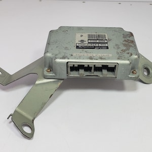 May include: A silver electronic control unit with a metal bracket. The rectangular unit has multiple connectors and a label with text and a barcode. The bracket has mounting holes. Likely for automotive use.