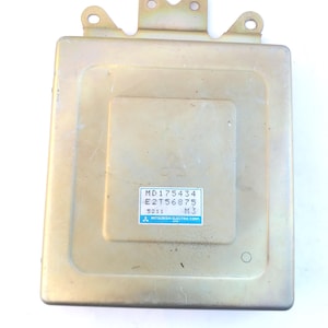 Puede incluir: Una pieza automotriz rectangular de color metálico con un soporte en la parte superior. Una etiqueta en la parte delantera muestra el texto "MD175434", "E2T56875", "5211" y "M3", junto con el logotipo "MITSUBISHI ELECTRIC CORP.".
