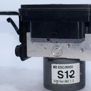 Op de afbeelding: Een zwart en zilver auto-onderdeel met een cilindrische basis en een rechthoekige bovenkant. Het onderdeel heeft een label met de tekst "MD ESC(RHD) S12 S/W Ver MD 1.3". Het heeft verschillende connectoren en montagebeugels.