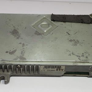 May include: A silver-coloured electronic control unit (ECU) with a rectangular shape. The ECU has a diamond-shaped indentation on the top and a barcode label. The unit has a series of fins on one side and a mounting bracket.
