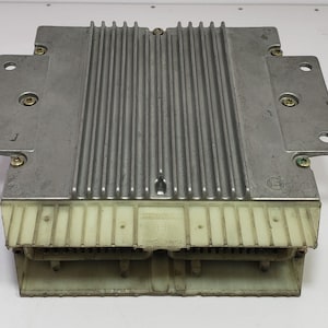 Puede incluir: Una unidad de control electrónico plateada con una parte superior acanalada y múltiples puntos de montaje. La unidad tiene forma rectangular con un conector beige claro en la parte inferior. Los tornillos aseguran los componentes. Es probable que sea una pieza de automóvil.