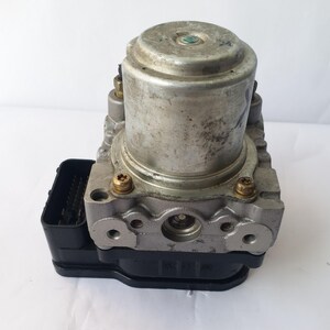 Op de afbeelding: Een zilverkleurige auto-ABS-module met een cilindrische bovenkant en een zwarte rechthoekige connector. De module heeft meerdere poorten en is vastgezet met goudkleurige schroeven. De unit is ontworpen voor voertuigremsystemen.
