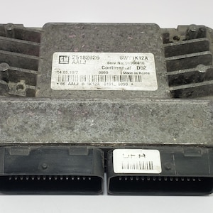 Puede incluir: Un módulo informático automotriz plateado con dos conectores negros y varias etiquetas. Las etiquetas incluyen texto y códigos de barras. El módulo tiene forma rectangular con una parte superior acanalada y orificios de montaje.